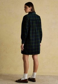 Kockovaná šaty v tmavej zelenej a námorskej farbe, s dlhými rukávmi, golierom a zadným dielom s nariasením. Nosené s ponožkami a mokasínami.