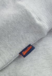 Pelēka sporta jakas ar mīkstu tekstūru, iekļaujot ribotās piedurknes un tumši zilā krāsā izstrādātu etiķeti ar uzrakstu "SuperDry" oranžā diegā.