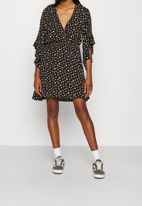 Schwarzes Wickelkleid mit einem floralen Muster in Gelb und Weiß, V-Ausschnitt und ausgestellt geschnittenen Ärmeln. Kombiniert mit grünen Turnschuhen und weißen Socken.