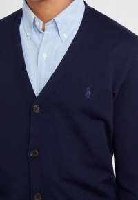 Námořnický modrý pletený kardigan s výstřihem do V, vybavený jedním knoflíkem na zapínání a malým vyšitým logem na levé hrudi.