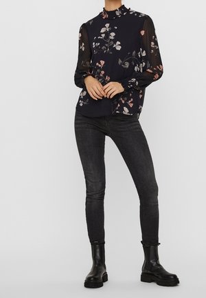 Personne portant un chemisier transparent noir à fleurs à manches longues, un jean skinny foncé et des bottines noires, debout les mains jointes devant.