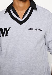 Persona che indossa un maglione grigio chiaro a V con rifiniture in bianco e nero sopra una camicia nera con colletto, caratterizzato da testo ricamato sul petto.