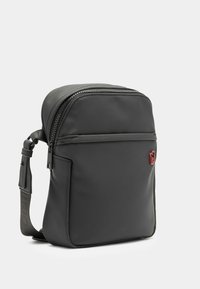 Musta olkalaukku, jossa on sileä, mattapintainen pinta, yksittäinen vetoketjutasku, säädettävä hihna ja pieni punainen logoelementti.