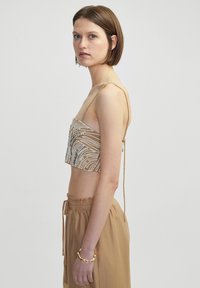 Kralen crop top van beige stof, met ingewikkelde witte patronen. Dunne verstelbare bandjes en een gestructureerde afwerking passen bij een lichtbruine onderkant.