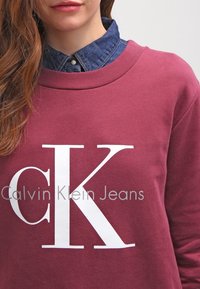 Sudadera burdeos con el logo blanco "CK" y el texto "Calvin Klein Jeans". Capa con cuello de mezclilla, con tela texturizada.