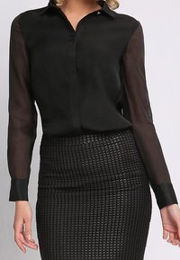 Blusa nera con maniche lunghe trasparenti, chiusura con bottoni e colletto; abbinata a una gonna a tubino nera con texture e un motivo geometrico.