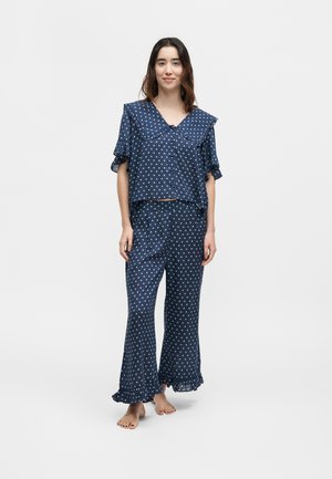 Γυναίκα που στέκεται ξυπόλητη φορώντας σετ πυτζάμες από σατέν σε σκούρο μπλε με πουά, με βολάν και φαρδιά γιακά και τελειώματα στα παντελόνια, σε απλό ανοιχτό φόντο.