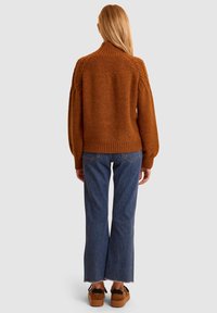 Maglione lavorato a maglia color ruggine con colletto alto, dettagli a treccia sulle spalle, abbinato a jeans blu svasati e scarpe con piattaforma marroni.