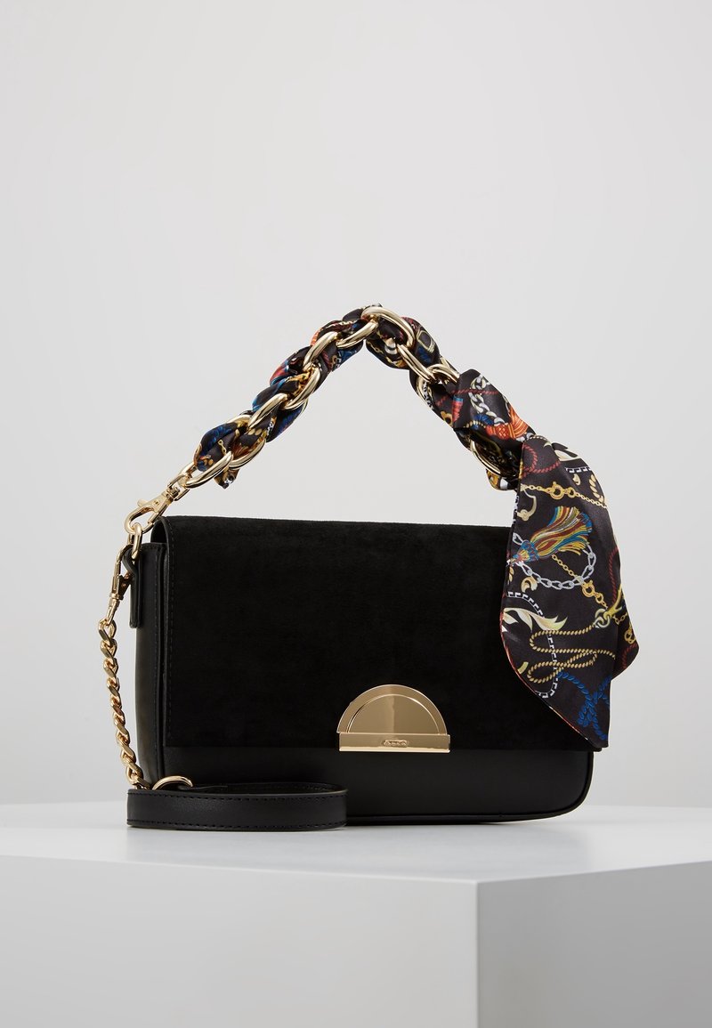 Sac à main noir avec fermoir doré, anse chaîne dorée enlacée d'une écharpe multicolore à motif, posé sur une surface blanche.