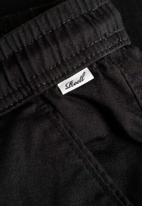 Primer plano de tela negra con cinturilla elástica y una etiqueta blanca de ropa con la palabra "Reell" cosida en la costura.