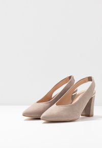 Beiga mockakälkar med blockklackar, spetsiga tår och öppna bakremmar på en vit yta med neutral bakgrund.