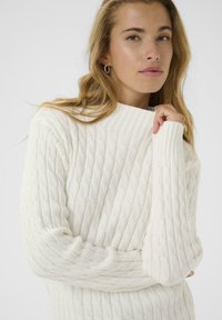 Maglione a lavorazione a trecce di colore bianco sporco, con collo alto e polsini a costine. Texture morbida con un motivo in rilievo per un effetto visivo interessante.