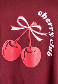 Bordeauxfarvet t-shirt med en grafik af to lyserøde kirsebær bundet med en hvid sløjfe, med teksten "cherry club" i en legesyg skrifttype.
