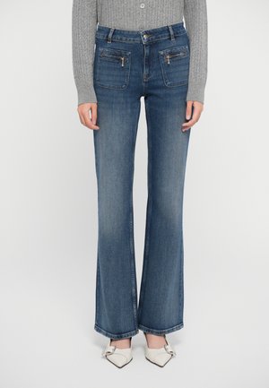 Jeans svasati blu scuro con due tasche frontali con zip, vita alta e orlo pulito, abbinati a scarpe bianche a punta e un maglione grigio a costine.