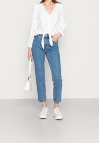 Femme portant un chemisier blanc noué devant, un jean taille haute bleu à petits pois blancs, des baskets blanches et tenant un sac à main blanc.
