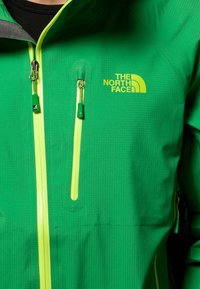 Grüne Jacke von The North Face mit strukturiertem Finish, gelben Akzenten, zwei Reißverschlüssen und einem gelben Logo auf der Vorderseite.