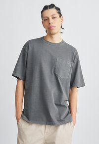 Szürke pamut rövid ujjú póló mellkasi zsebbel. Laza szabással és kerek nyakkivágással rendelkezik. Texturált, kopott hatású felület.