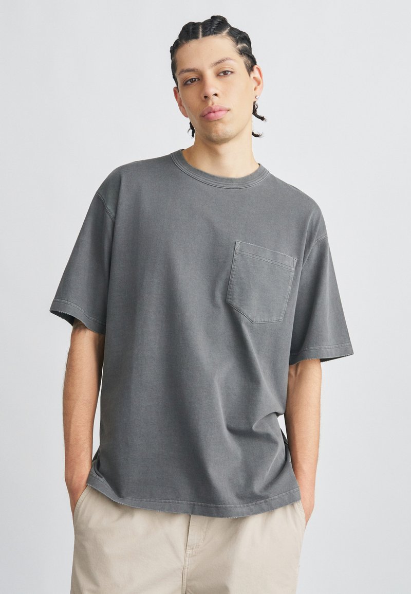 Szürke pamut rövid ujjú póló mellkasi zsebbel. Laza szabással és kerek nyakkivágással rendelkezik. Texturált, kopott hatású felület.