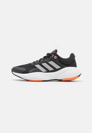 Zwarte sportschoen met grijze strepen, roze veterogen, dikke witte zool en oranje loopvlakaccenten op een effen witte achtergrond.