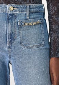 Vaqueros de mezclilla de color azul claro con una cintura alta, que presentan un detalle de cadena dorada en el bolsillo trasero y un ligero desvanecimiento para aportar textura.