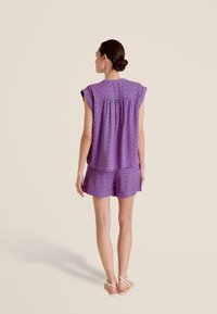 Ensemble court à motifs violets avec manches courtes, présentant une coupe ample et un détail froncé au dos, associé à des sandales à lanières.