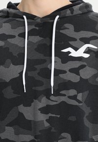 Hoodie i svart och grå camouflage-mönster, med vita dragsko och en vit logotyp på bröstet. Mjuk tygstruktur.