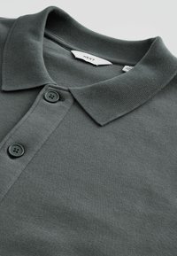 Polo gris foncé avec deux boutons et un col côtelé, montrant une étiquette de marque blanche « NEXT » à l'intérieur de l'encolure.