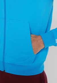 Blaues Zip-Jackett aus strukturiertem Stoff mit einer Vorderentasche. Bündchen und Saum in einheitlichem Blau. Sichtbare Hand in der Tasche.