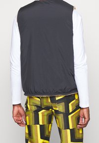 Svart vadderad väst med en mjuk beige krage, bärs över en vit långärmad skjorta, ihop med byxor med gult och grönt geometriskt mönster.