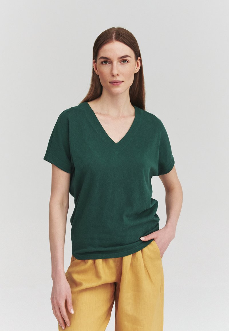 Femme aux longs cheveux bruns portant un haut vert foncé à manches courtes en V et un pantalon jaune moutarde, debout avec une main dans la poche.