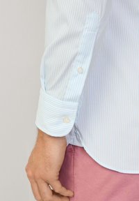 Camisa a rayas azul claro con puños abotonados, que presenta una textura suave y pequeños botones blancos. La tela tiene un ligero brillo.