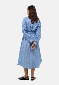 Robe longue à rayures bleu clair avec une taille ceinturée, des manches longues et atteignant le mi-mollet. Des sandales plates noires complètent le look.