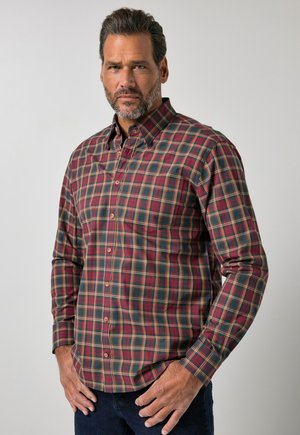 Homme avec barbe portant une chemise à carreaux rouge et verte boutonnée, debout devant un fond gris clair uni.