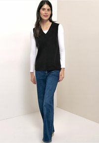 Zwarte mouwloze V-halsvest over een witte top met lange mouwen, gecombineerd met blauwe wijde jeans. Gladde textuur en een ontspannen pasvorm.