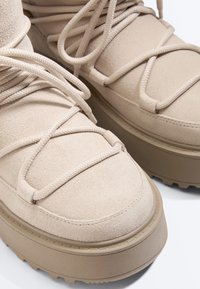 Bottes compensées en daim beige avec semelles épaisses texturées et lacets ronds assortis croisés sur le devant.