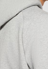 Sudadera gris con capucha de tejido suave y texturizado. Incluye una capucha redondeada y una vista en primer plano del detalle de la costura a lo largo del hombro.