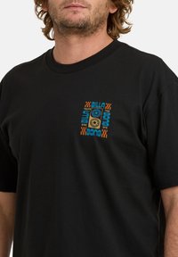 Mężczyzna w czarnej koszulce z małą kolorową grafiką i napisem "Billabong since 1973" na lewej piersi.