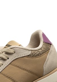 Beige sneakers med en strukturerad ljusbrun panel, ljusgrå accenter och en lila detalj vid hälen. Snörningsdesign med vadderad krage.