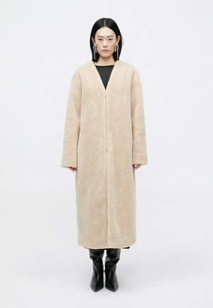 Long, beige, fluffy coat made from textured material, featuring a deep V-neck and wide sleeves, worn over a black top and paired with black boots.