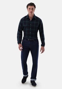 Camicia a quadri verde scuro e blu navy, abbinata a jeans blu scuro con le maniche arrotolate, indossata con scarpe in scamosciato nere.