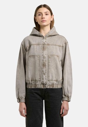 Vrouw draagt een lichtgrijs zip-up capuchonjack met voorzakken en elastische boorden, gecombineerd met een zwarte broek, staand tegen een effen achtergrond.