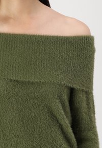 Maglione verde oliva con scollo a barchetta realizzato in un materiale soffice e peloso, con texture a costine sul collo e vestibilità ampia, che mette in risalto le spalle.
