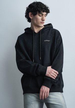 Młody mężczyzna o kręconych włosach, ubrany w czarną bluzę Off-White z delikatnymi nakrapianiami, poprawiający rękaw, stojący na tle jednolitego szarego tła.