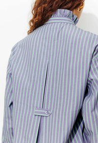 Blouse rayée violette et verte avec un col à volants haut, une fente au dos et un détail de boutons à la taille. Texture de tissu lisse.