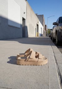 Sandales à plateforme beiges avec deux brides sur un trottoir en ville à côté de voitures garées et de bâtiments industriels sous un ciel bleu clair.