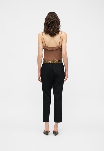Femme aux cheveux bruns bouclés portant un haut marron sans manches, un pantalon noir avec une ceinture imprimée léopard et des sandales noires à talons, vue de dos.