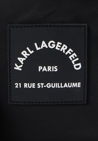 Μαύρη υφασμάτινη ετικέτα με ανάγλυφο λευκό κείμενο. Το κείμενο περιλαμβάνει "KARL LAGERFELD", "ΠΑΡΙΣΙ", και "21 RUE ST-GUILLAUME". Στρογγυλός σχεδιασμός με ραμμένο περίγραμμα.