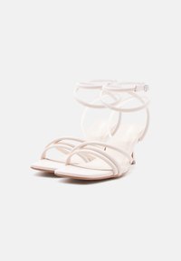 Hvide strop-sandaler med høje hæle, tynde ankelstropper og åbne firkantede tåpartier, placeret mod en ensartet hvid baggrund.