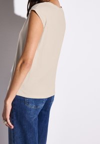 Top beige a maniche corte realizzato in morbido cotone, abbinato a jeans in denim blu. Il top presenta un collo rotondo e una vestibilità rilassata.
