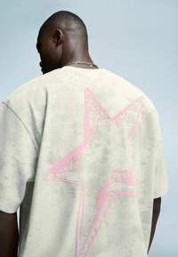 Man wearing light gray oversized t-shirt with textured pink star graphic on the back, facing away against plain light blue background.